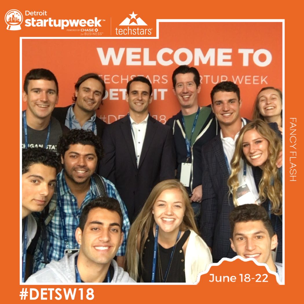 A large group poses for picture in the photo booth in front of the custom step and repeat backdrop for Detroit Startup Week
